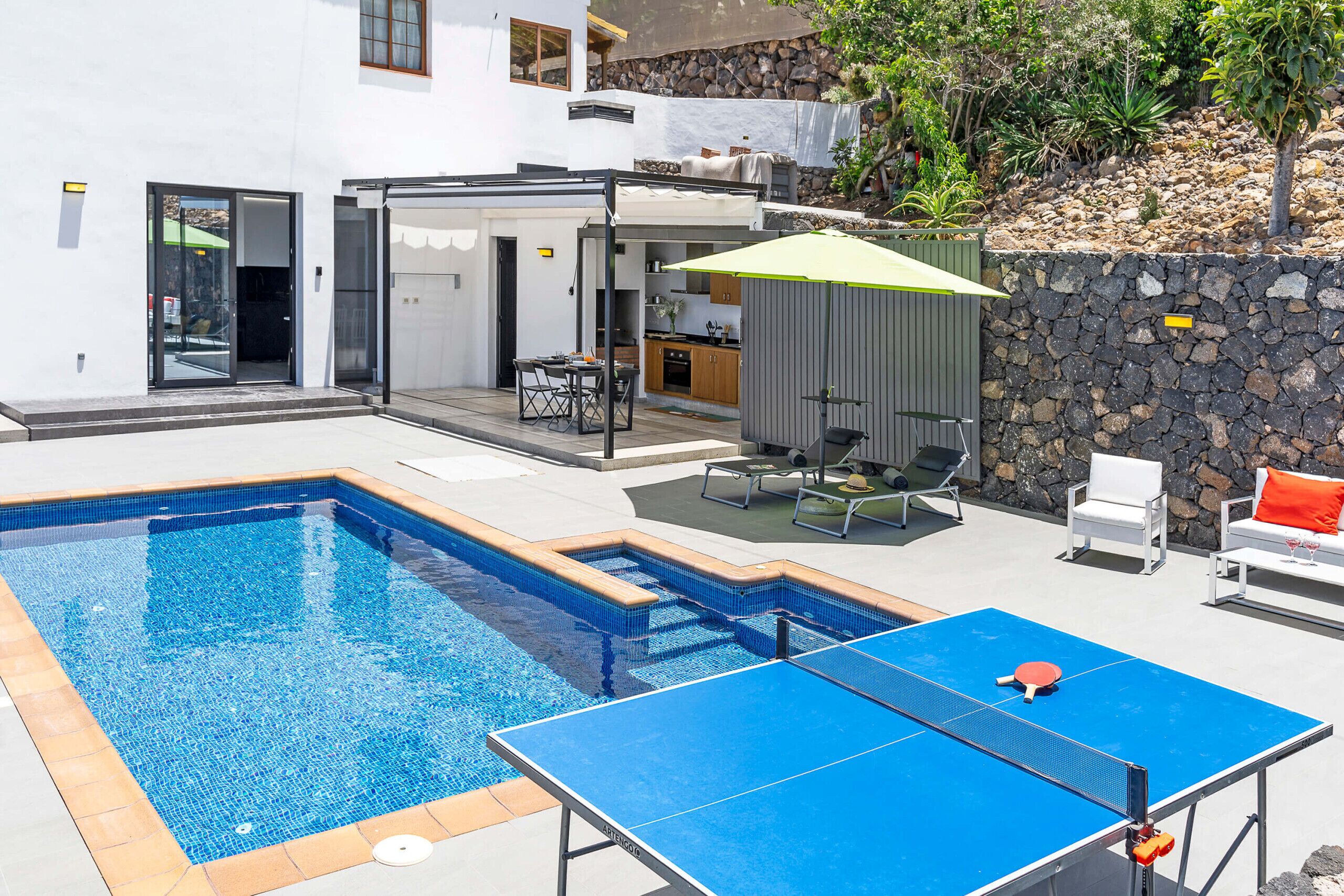 A swimming pool with a ping pong table, outdoor seating, and a kitchen area.