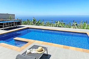 A swimming pool with a view of the ocean, a lounge chair, and a table with a hat and magazine. - Casa Juliana Teresa (Tijarafe)