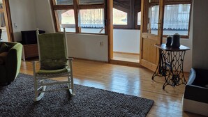 Una habitación con piso de madera, una silla verde, una mesita con dos jarrones y un ventanal con vistas al exterior.