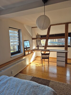 Quarto com teto inclinado, escrivaninha, cadeira, radiador e janela com persianas.
