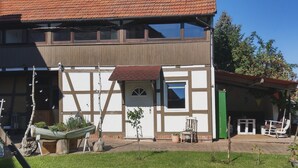 Un edificio de dos pisos con techo de tejas, un balcón y un pequeño jardín con césped y plantas en macetas.
