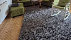 Una sala de estar acogedora con un sofá verde, un pequeño reposapiés, un sillón mecededor, una mesa de centro de madera y una alfombra estampada.