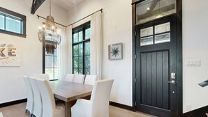 Een eetruimte met een houten tafel en witte stoelen, een kroonluchter, en een zwarte deur met een raam.