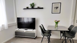 A modern living room with a flat-screen TV on a white stand, a dining table with four chairs, and a small potted plant on the floor.