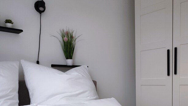 A neatly made bed with white linens and pillows, a black bedside table with a plant, and a white wardrobe with two doors.