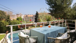 A poolside area with wicker chairs and a table, surrounded by greenery and a view of buildings.