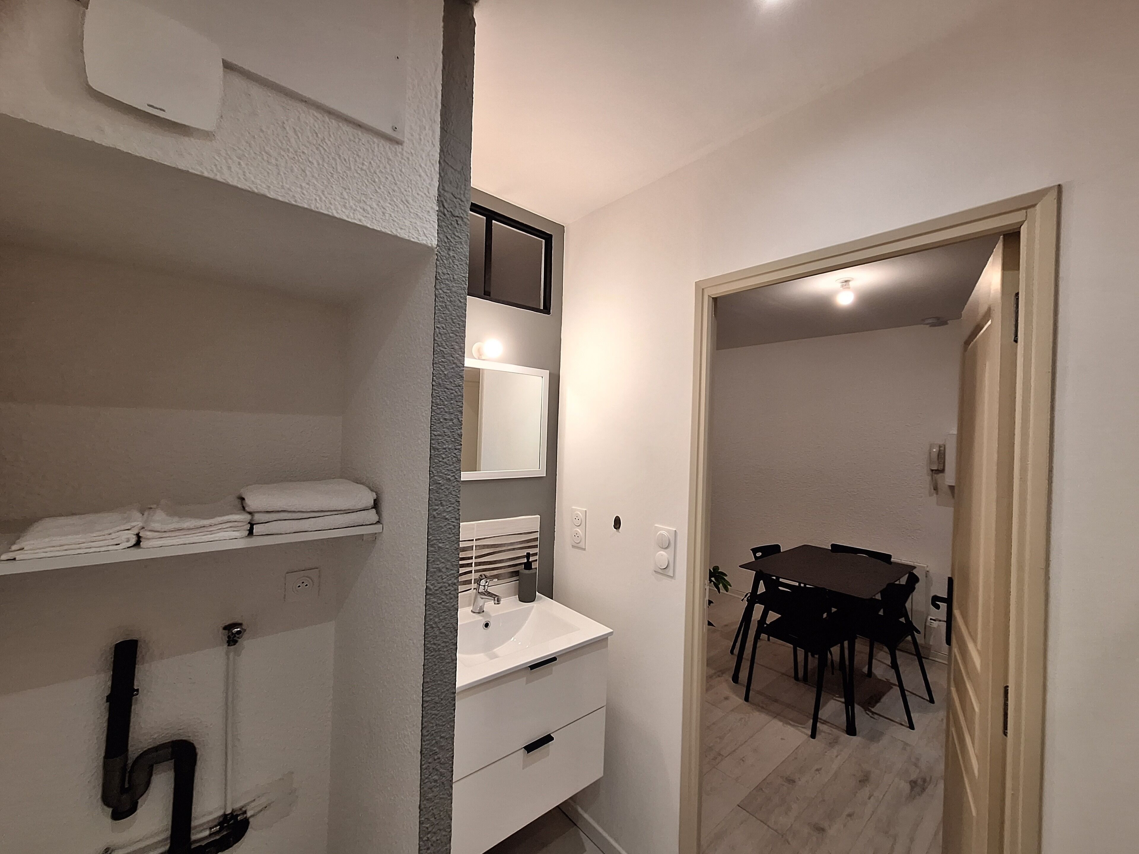 A bathroom with a sink, mirror, and towel rack. There is a door leading to a dining area with a table and chairs.