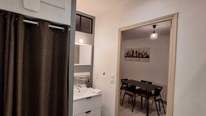A bathroom with a shower curtain, a sink, and a mirror. There is a dining area with a table and chairs. - Attractive, renovated 2-room apartment by the lake (Saint-Jean-d'Angély)