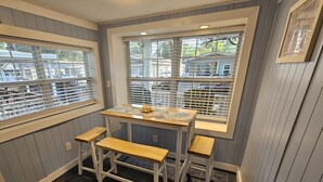 Quarto de canto com duas janelas, uma mesa com toalha estampada e três banquinhos de madeira.