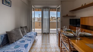 Une pièce comprenant un canapé, une table à manger et des chaises, un balcon avec des rideaux et un téléviseur posé sur un meuble en bois.