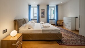 Une chambre à coucher avec un lit, une table de chevet avec une lampe, une chaise, une commode et une fenêtre avec des rideaux.
