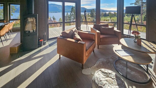 Amplia sala de estar con chimenea, asientos cómodos y ventanas grandes con vistas a un lago y montañas.
