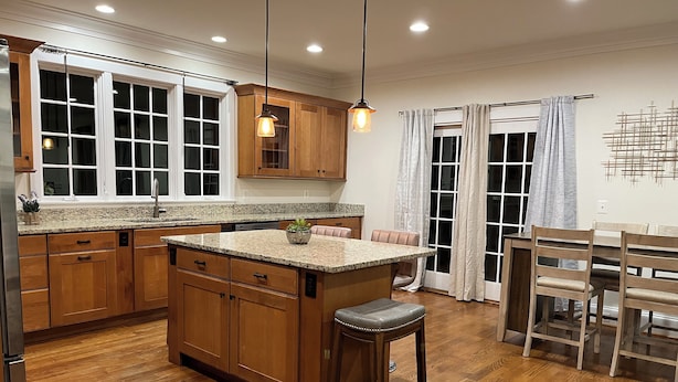 Una cocina con armarios de madera, encimeras de granito, una isla central con un taburete, iluminación colgante y una zona de comedor con mesa y sillas.