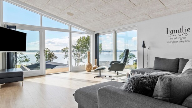 Ein geräumiges Wohnzimmer mit großem Fenster mit Blick auf ein Gewässer, einem grauen Sofa, einem schwarzen Sessel und einem an der Wand befestigten Flachbildfernseher.