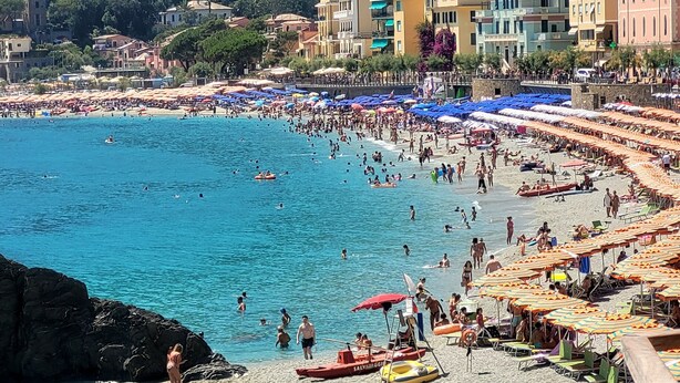 Una playa concurrida, gente nadando y tomando el sol, rodeada de edificios y sombrillas.