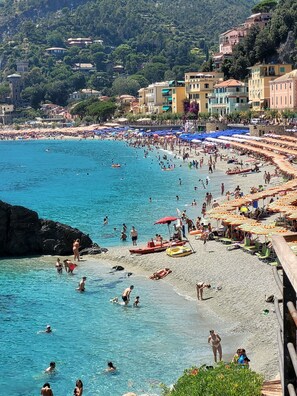 A crowded beach with people swimming and sunbathing, surrounded by buildings and umbrellas.