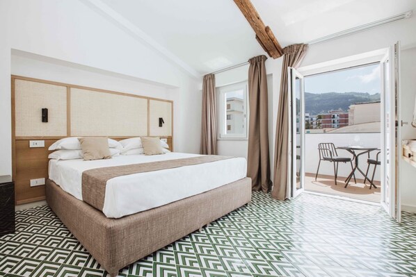 A hotel room with a large bed, a wooden headboard, a balcony with a table and chairs, and patterned flooring.
