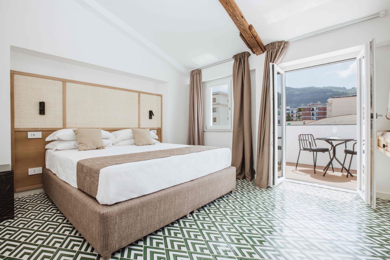 A hotel room with a large bed, a wooden headboard, a balcony with a table and chairs, and patterned flooring.