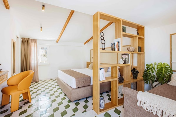A hotel room with a bed, a wooden bookshelf, a desk, an orange chair, and a potted plant.
