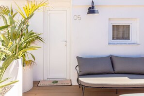 A room with a white door, a sofa with grey cushions, a potted plant, a wall-mounted lamp, and a window with blinds.