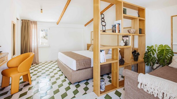 Een hotelkamer met een bed, een houten boekenkast, een bureau, een oranje fauteuil en een potplant.