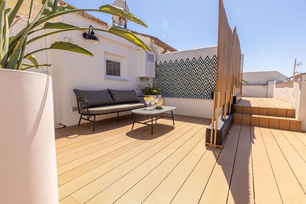 A wooden deck with a sofa and a table, surrounded by a white wall with a geometric pattern.