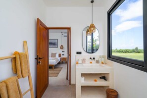 A modern bathroom with a large mirror, a sink, and a window with a view of greenery. - Villa Flambard (Canggu)