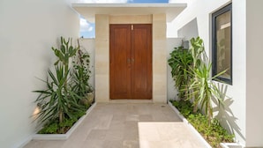 A modern building entrance with a wooden double door, surrounded by potted plants and a tiled pathway. - Villa Flambard (Canggu)
