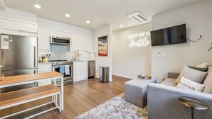 A modern kitchen with stainless steel appliances, a dining area with a wooden table, and a living room with a grey sofa and a flat-screen TV mounted on the wall. - Travelers Dream-Steps to Space Needle- Shared Backyard (Seattle)