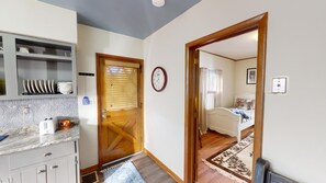 A compact room with a wooden door, a clock, a bed, and a small kitchen area with a sink and cabinets.
