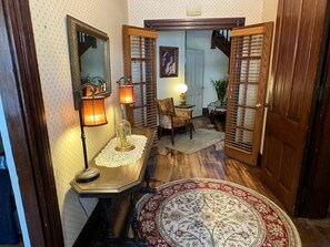 A hallway with a wooden floor, a round patterned rug, a table with a lamp, a chair, and a mirror.