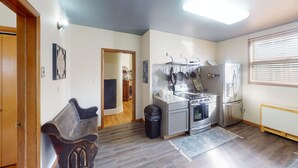 A compact kitchen with a stove, oven, and refrigerator. There is a wooden bench and a trash bin.