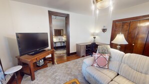 A living room with a television on a wooden stand, a sofa, a desk with a chair, and a wooden wardrobe.