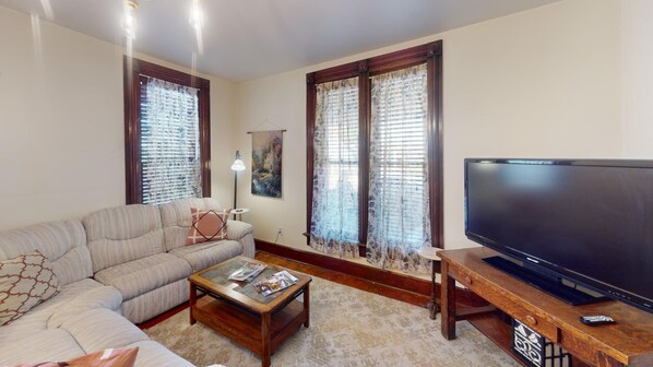 Un salón con sofá, mesa de centro, televisor y ventanas con cortinas.