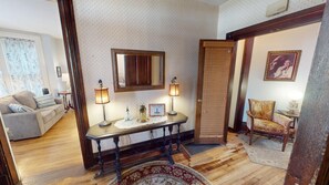 Una habitación tradicional con suelo de madera, un sofá, una mesa con lámparas, un espejo y una puerta con marco de madera.