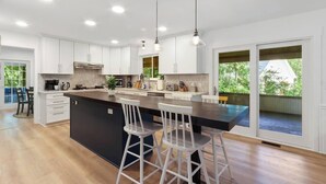 A modern kitchen with white cabinets, a large island with bar stools, and a view of the outdoors through sliding glass doors. - Wildwood Retreat - Theater, Hot Tub and More! (Lake Geneva)