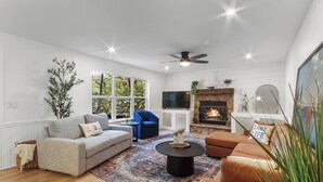 A modern living room with a fireplace, a ceiling fan, and a large window overlooking greenery. - Wildwood Retreat - Theater, Hot Tub and More! (Lake Geneva)