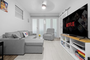 A modern living room with a grey sectional sofa, a grey armchair, a flat-screen TV, and a bookshelf with books and games.