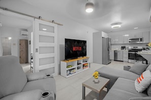 A modern living room with a flat-screen TV, grey sofas, a coffee table, and a kitchen in the background.