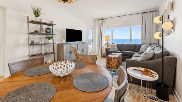 Un salón moderno con una mesa de comedor de madera, cojines grises y un gran ventanal con vistas al mar.
