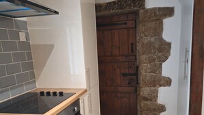 Eine Küche mit Herd, Backofen und Schränken. Im Hintergrund ist eine Steinwand und eine Holztür zu sehen.