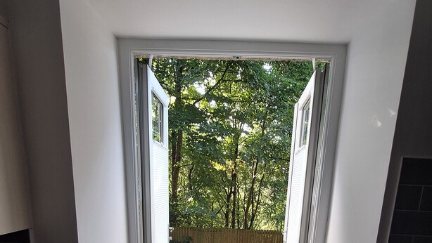 Blick vom Eingangsbereich eines Zimmers auf einen Balkon mit Holzboden, Gartenmöbeln und einem Metallgeländer.