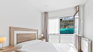Un dormitorio con cama, mesita de noche, lámpara, ventana con cortinas y vistas al exterior.