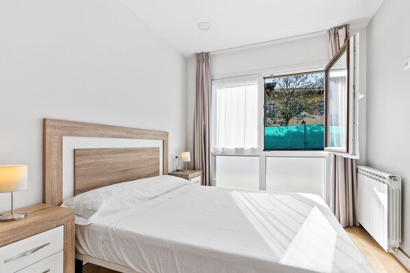 A bedroom with a bed, bedside table, lamp, window with curtains, and a view of greenery outside.