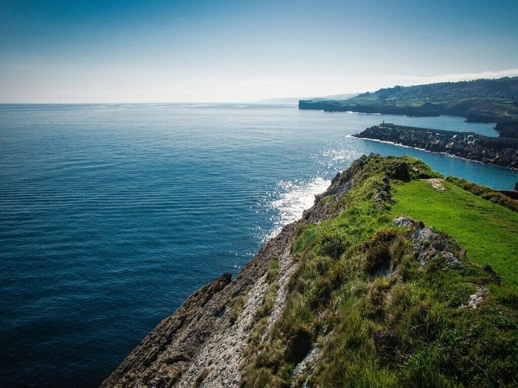 A coastal landscape with a cliff overlooking the sea, a grassy area, and distant hills.