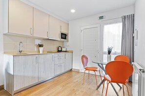 A modern kitchen with a microwave, sink, and cabinets. There is a dining area with a glass table and orange chairs. A sign above the door reads 'SALIDA.'