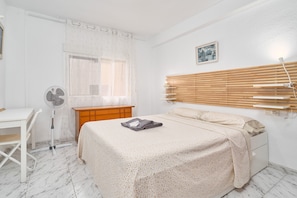 Een slaapkamer met een bed, een bureau, een stoel, een nachtkastje, een lamp, een raam met zonwering en een ventilator.
