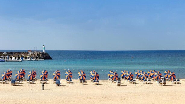 Een strand met rijen strandstoelen en parasols, een vuurtoren en boten op de achtergrond.