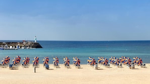 Une plage bordée de rangées de chaises longues et de parasols, un phare et des bateaux en arrière-plan.