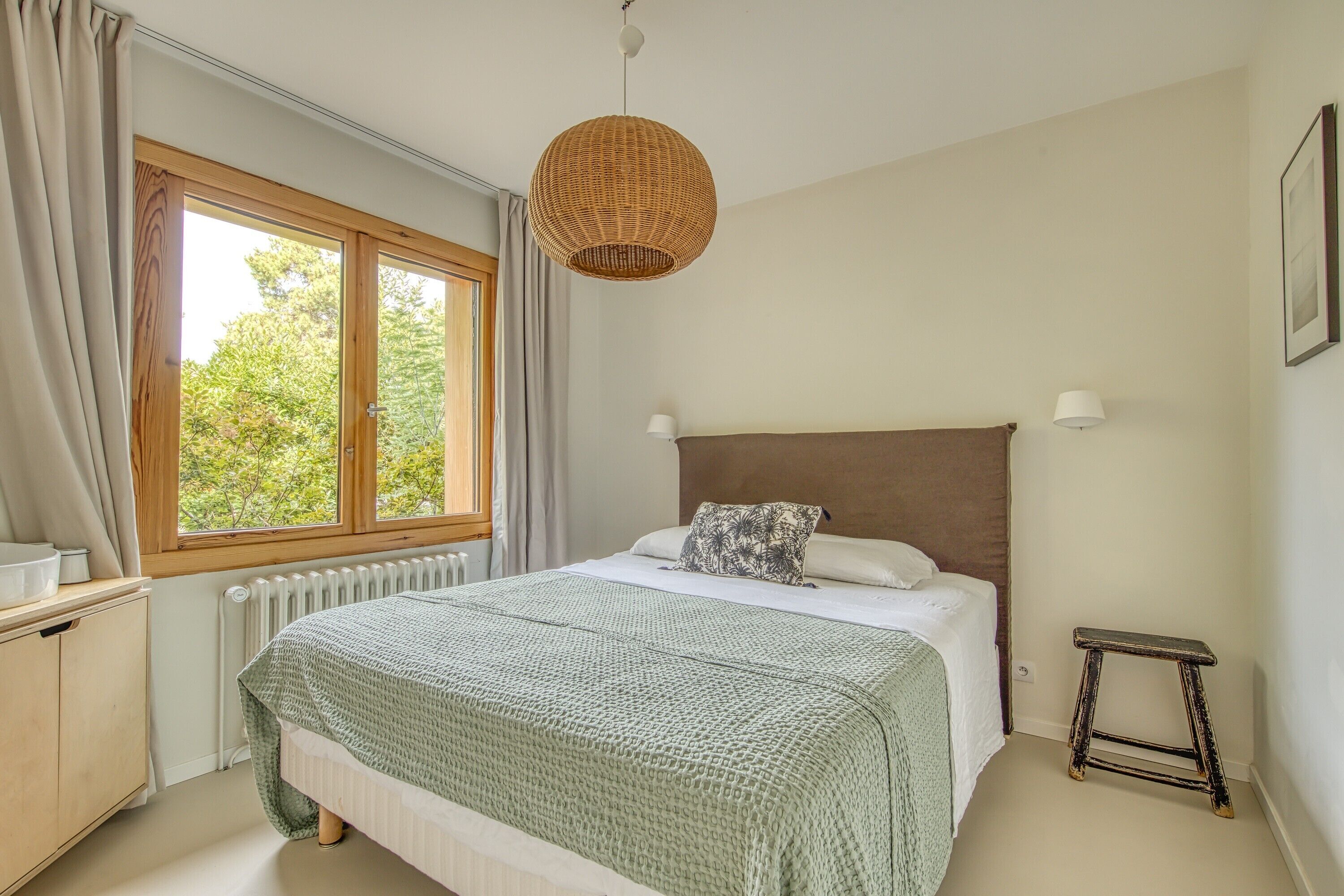 A bedroom with a large window, a bed with a green blanket, a wooden headboard, a wicker hanging light, and a small stool.